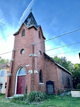 A red brick church with pointed arched windows and a tall steeple with a cross on top. The structure includes white stone accents and a large wooden door. The sky is clear and blue, while some greenery is visible around the building.