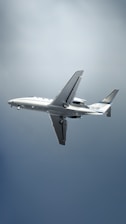 A white commercial airplane is in mid-flight against a backdrop of a clear sky. Landing gear is partially extended. The plane features blue and white stripe accents near the tail with registration markings visible.