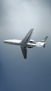 A white commercial airplane is in mid-flight against a backdrop of a clear sky. Landing gear is partially extended. The plane features blue and white stripe accents near the tail with registration markings visible.