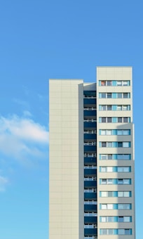 A modern high-rise building featuring multiple floors with large windows. The facade is primarily light gray, accented with dark blue panels. The sky is clear and bright, contributing to a minimalist aesthetic.