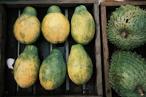 A wooden crate containing six partially ripe papayas with a mix of green and yellow skin arranged neatly. Adjacent to this crate is another box with two large green soursops featuring a bumpy texture.