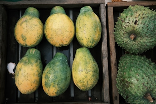A wooden crate containing six partially ripe papayas with a mix of green and yellow skin arranged neatly. Adjacent to this crate is another box with two large green soursops featuring a bumpy texture.