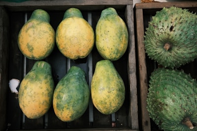 A wooden crate containing six partially ripe papayas with a mix of green and yellow skin arranged neatly. Adjacent to this crate is another box with two large green soursops featuring a bumpy texture.