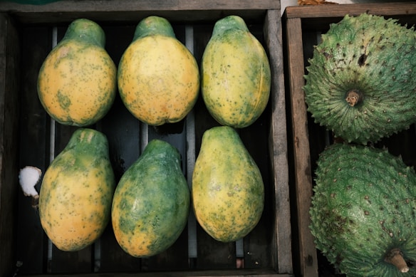 A wooden crate containing six partially ripe papayas with a mix of green and yellow skin arranged neatly. Adjacent to this crate is another box with two large green soursops featuring a bumpy texture.