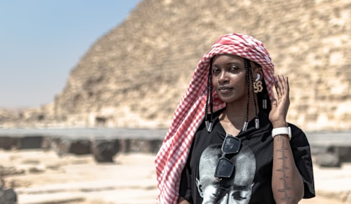 A person wearing a red and white checkered headscarf stands in front of a pyramid. The individual is dressed in a black t-shirt with a portrait on it and has sunglasses hanging from the collar. They have earphones in their ears and wear a white smartwatch on their wrist. The background is dominated by the blurred image of the stone blocks of the pyramid.