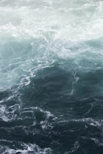 A swirling, dynamic ocean scene with shades of blue and green. The water appears rough with visible waves and currents creating intricate patterns across the surface.
