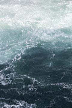 A swirling, dynamic ocean scene with shades of blue and green. The water appears rough with visible waves and currents creating intricate patterns across the surface.