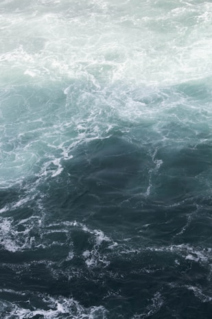 A swirling, dynamic ocean scene with shades of blue and green. The water appears rough with visible waves and currents creating intricate patterns across the surface.