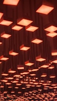 A ceiling installation featuring numerous square light fixtures, each emitting a warm, orange glow. The lights are arranged in a grid pattern and are suspended at varying heights, creating a visually dynamic effect.