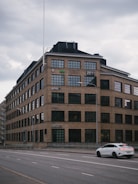 A large, multi-story brick building with numerous rectangular windows. The building features company logos on its facade and a digital sign reading '2011'. A white car is passing by on a wide, empty road in front of the building. The sky is overcast, giving a somber appearance.