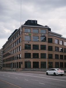 A large, multi-story brick building with numerous rectangular windows. The building features company logos on its facade and a digital sign reading '2011'. A white car is passing by on a wide, empty road in front of the building. The sky is overcast, giving a somber appearance.