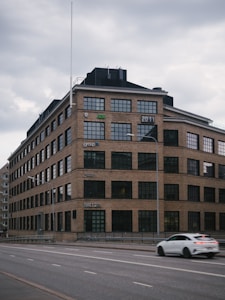 A large, multi-story brick building with numerous rectangular windows. The building features company logos on its facade and a digital sign reading '2011'. A white car is passing by on a wide, empty road in front of the building. The sky is overcast, giving a somber appearance.