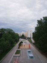 A basketball court and ping pong tables are situated along a paved walkway lined with tall, green trees and well-maintained hedges. High-rise residential buildings with light-colored facades form the background beneath a cloudy sky.