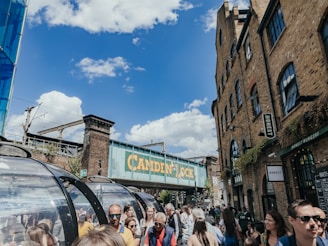 A bustling urban scene featuring a crowd of people in a market area under a clear blue sky. A bridge labeled 'Camden Lock' spans overhead, bordered by historic brick buildings with signage for eateries and shops. The atmosphere is lively and vibrant, conveying a sense of a popular, tourist-friendly area.