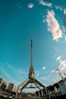 A large, industrial crane dominates the foreground, set against a vivid blue sky with scattered clouds. Residential buildings and a ship are visible in the background, with details suggesting a harbor or dockside environment.