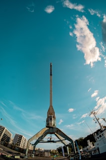 A large, industrial crane dominates the foreground, set against a vivid blue sky with scattered clouds. Residential buildings and a ship are visible in the background, with details suggesting a harbor or dockside environment.