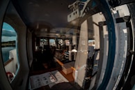 A cabin interior with large windows, reflecting parts of a boat structure and showing the outdoor landscape featuring water and trees. Inside the cabin are various nautical instruments, a computer monitor, and seating, indicating a control room or bridge on a vessel.
