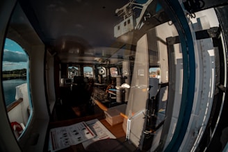 A cabin interior with large windows, reflecting parts of a boat structure and showing the outdoor landscape featuring water and trees. Inside the cabin are various nautical instruments, a computer monitor, and seating, indicating a control room or bridge on a vessel.