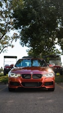 A red BMW car is prominently parked on a road with its headlights on, flanked by golf carts in the background. Large trees with dense foliage provide shade behind the vehicle, while the road and grass hint at a park or leisure environment.