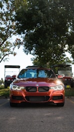 A red BMW car is prominently parked on a road with its headlights on, flanked by golf carts in the background. Large trees with dense foliage provide shade behind the vehicle, while the road and grass hint at a park or leisure environment.