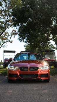 A red BMW car is prominently parked on a road with its headlights on, flanked by golf carts in the background. Large trees with dense foliage provide shade behind the vehicle, while the road and grass hint at a park or leisure environment.
