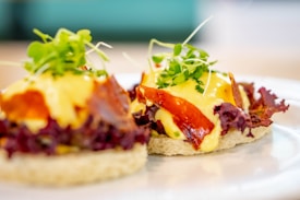 A close-up view of two gourmet appetizers featuring toasted bread topped with creamy yellow sauce, crispy bacon, fresh green sprouts, and purple lettuce, all elegantly arranged.