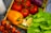 A variety of fresh vegetables and fruits are arranged in a wooden crate. There are several red tomatoes on the vine, an orange bell pepper, a purple eggplant, green chilies, a half-cut lemon, and green leafy lettuce.