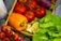 A variety of fresh vegetables and fruits are arranged in a wooden crate. There are several red tomatoes on the vine, an orange bell pepper, a purple eggplant, green chilies, a half-cut lemon, and green leafy lettuce.