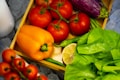 A variety of fresh vegetables and fruits are arranged in a wooden crate. There are several red tomatoes on the vine, an orange bell pepper, a purple eggplant, green chilies, a half-cut lemon, and green leafy lettuce.