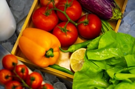 A variety of fresh vegetables and fruits are arranged in a wooden crate. There are several red tomatoes on the vine, an orange bell pepper, a purple eggplant, green chilies, a half-cut lemon, and green leafy lettuce.