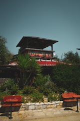 A rustic outdoor scene featuring a two-story structure with wooden elements and a red tiled roof. The upper level is open, supported by wooden beams, and adorned with colorful shapes. Lush green foliage and various types of plants are in the foreground, along with stonework and a few red wooden benches facing the structure. The sky is a muted teal color.