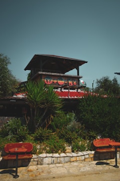 A rustic outdoor scene featuring a two-story structure with wooden elements and a red tiled roof. The upper level is open, supported by wooden beams, and adorned with colorful shapes. Lush green foliage and various types of plants are in the foreground, along with stonework and a few red wooden benches facing the structure. The sky is a muted teal color.