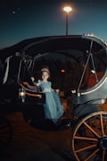 A young child, dressed as a princess complete with a tiara, sits elegantly in a vintage horse-drawn carriage at night. The child is wearing a light blue gown, and a warm lamp illuminates the scene from the carriage’s side. A tall streetlamp in the background casts a soft glow over the surroundings, adding a magical atmosphere.