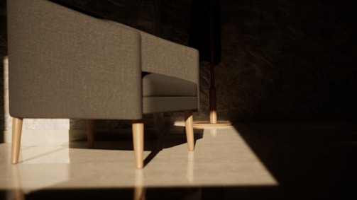 A modern gray sofa with light wooden legs sits in a room with minimalistic decor. Sunlight creates a dramatic pattern on the polished floor, highlighting the clean lines of the furniture. The background is dark, with a subtle texture on the walls, which enhances the contrast between the illuminated area and the shadows.