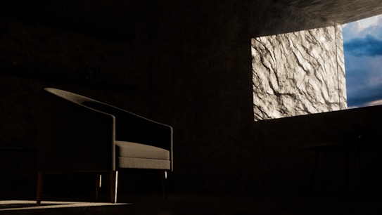 A dimly lit room featuring a dark armchair positioned towards the left, with a textured wall on the right illuminated by daylight streaming through a window. The window offers a partial view of a cloudy blue sky.