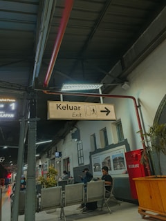 A station platform with several people sitting and standing, some facing away from the camera. There is a sign indicating the direction of the exit with the word 'Keluar' which is 'Exit' in English and an arrow pointing right. The ceiling has exposed metal beams, and there are plants and seating areas visible. A red fire hydrant is affixed to the wall, and a pole supporting the exit sign extends from the ceiling.