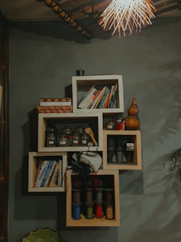 A creatively designed wall shelf with an arrangement of books on the top and middle sections. Several glass jars containing various items are neatly placed in one section. Colorful spools of thread in multiple compartments add vibrancy. A decorative light fixture with a woven design hangs from the ceiling, casting a warm glow over the setting. The overall ambiance is cozy and eclectic.