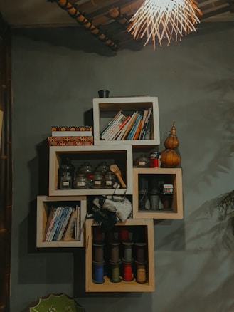 A creatively designed wall shelf with an arrangement of books on the top and middle sections. Several glass jars containing various items are neatly placed in one section. Colorful spools of thread in multiple compartments add vibrancy. A decorative light fixture with a woven design hangs from the ceiling, casting a warm glow over the setting. The overall ambiance is cozy and eclectic.