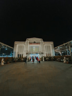 A large building marked with the name 'Yogyakarta' at the top is set against a dark night sky. The structure is illuminated, showcasing its architectural details. Several people are seen entering or exiting the building, some pulling luggage. Potted plants line the path leading up to the entrance.