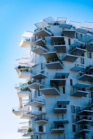 A modern, high-rise building with an innovative design featuring numerous protruding balconies. Each balcony is adorned with greenery, adding a touch of nature to the architectural structure. The building appears to be white with blue shutters, contrasting against the clear blue sky. Angular shapes and overhangs give a dynamic and futuristic look.