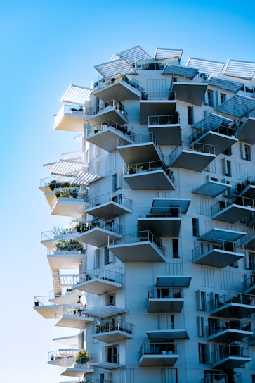 A modern, high-rise building with an innovative design featuring numerous protruding balconies. Each balcony is adorned with greenery, adding a touch of nature to the architectural structure. The building appears to be white with blue shutters, contrasting against the clear blue sky. Angular shapes and overhangs give a dynamic and futuristic look.