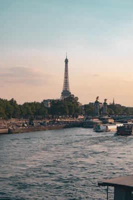 The Eiffel Tower stands prominently in the background as the sun sets, casting a warm glow over the scene. The Seine River flows gently, lined with trees and populated with boats and people enjoying the scenic view. The sky transitions from a gentle blue to a soft orange, enhancing the tranquil atmosphere.