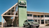 A Land Rover dealership building with a green Land Rover Defender parked outside under a clear blue sky. The dealership sign displays 'Land Rover Redwood City' in white text against a green background. The architecture features a modern design with large windows and a prominent geometric roof structure.