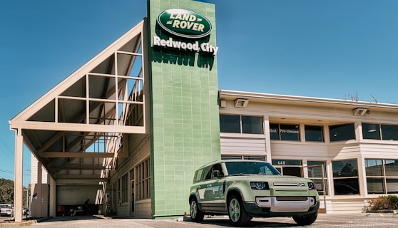 A Land Rover dealership building with a green Land Rover Defender parked outside under a clear blue sky. The dealership sign displays 'Land Rover Redwood City' in white text against a green background. The architecture features a modern design with large windows and a prominent geometric roof structure.