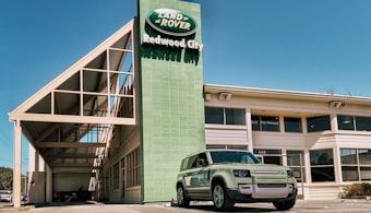 A Land Rover dealership building with a green Land Rover Defender parked outside under a clear blue sky. The dealership sign displays 'Land Rover Redwood City' in white text against a green background. The architecture features a modern design with large windows and a prominent geometric roof structure.