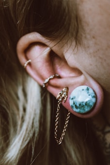 A close-up view of an ear adorned with multiple pieces of jewelry, including a large stone plug and several gold rings with chains. The stone plug has a greenish marble texture.