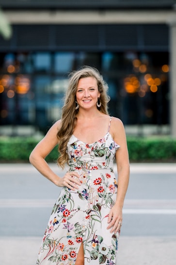 A woman with long hair stands outdoors, wearing a floral-patterned dress. She has a confident smile and is posing with one hand on her hip. The background features blurred lights and a building facade, suggesting an urban setting.