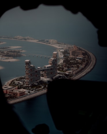 An aerial view of a man-made island shaped like a palm tree, featuring large, luxurious buildings and hotels along the trunk. The water surrounding the island is a deep blue, and the infrastructure includes roads and smaller structures.