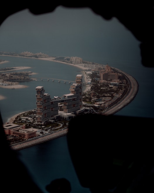 An aerial view of a man-made island shaped like a palm tree, featuring large, luxurious buildings and hotels along the trunk. The water surrounding the island is a deep blue, and the infrastructure includes roads and smaller structures.