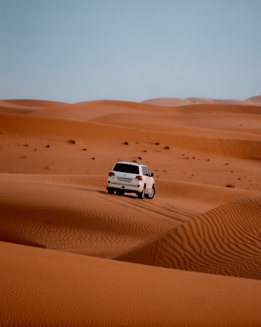 A white SUV is driving over vast orange sand dunes under a clear blue sky. The tracks of the vehicle are visible on the sand, suggesting movement and adventure.